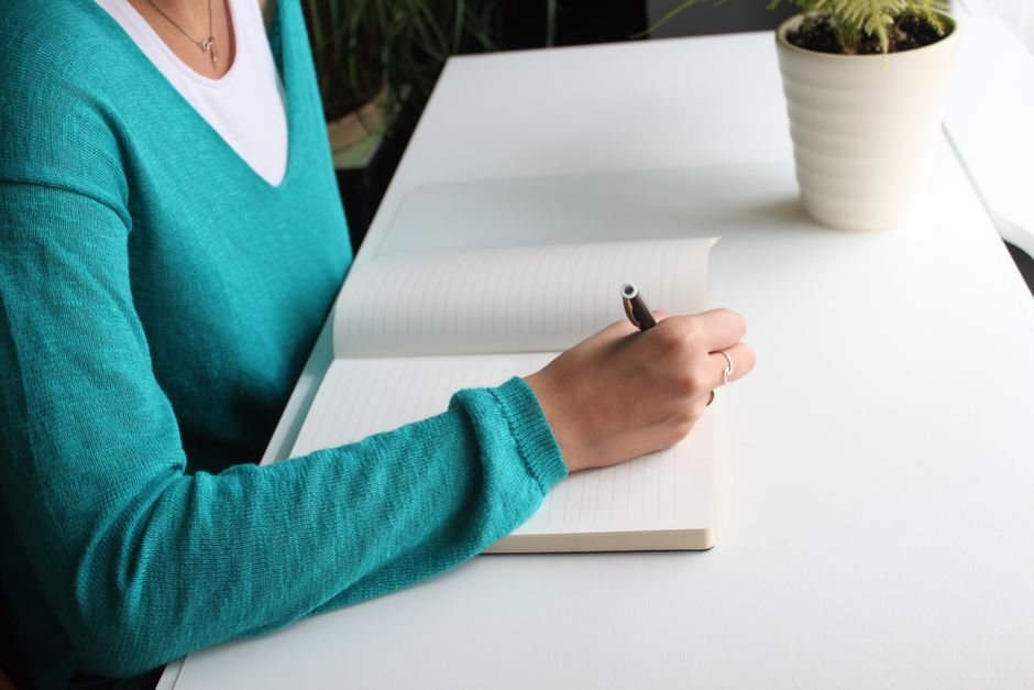 Pessoa anotando ideias em um caderno branco, em um ambiente de trabalho organizado com plantas ao fundo.