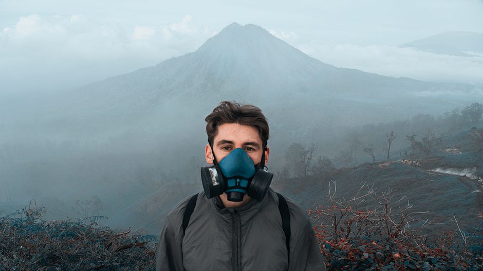 Homem usando máscara de proteção em meio a uma paisagem de vulcão e natureza, simbolizando cuidados ambientais ou prevenção de riscos em áreas de risco.