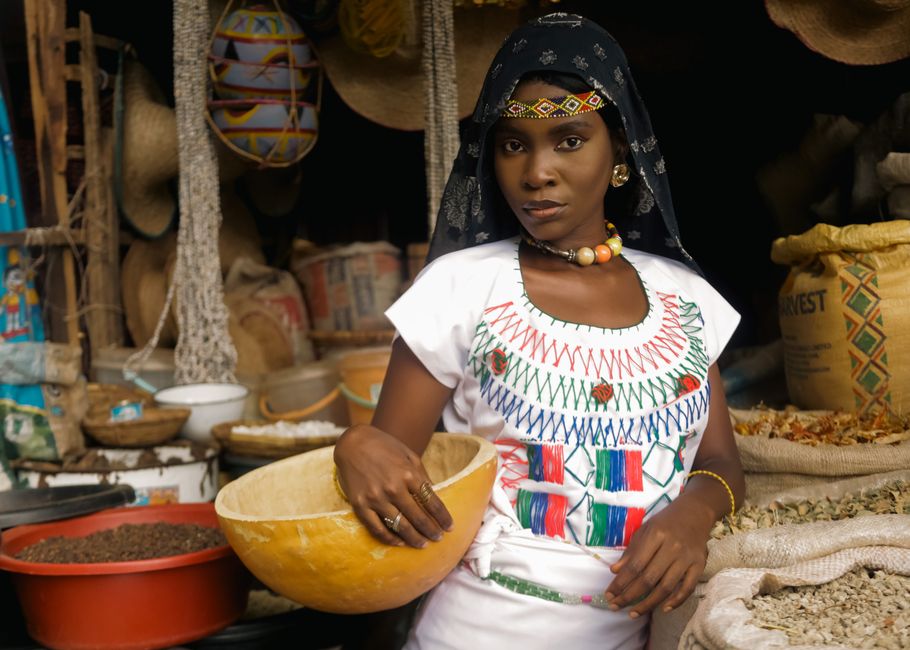Mulher africana em mercado tradicional, vestida com traje cultural e rodeada de produtos naturais e artesanais.