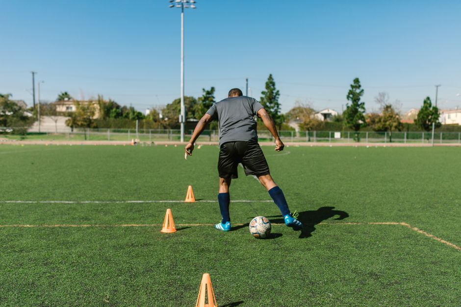 Homem treinando habilidades de controle de bola em um campo de futebol ao ar livre, usando cones para treino de dribles e coordenação.