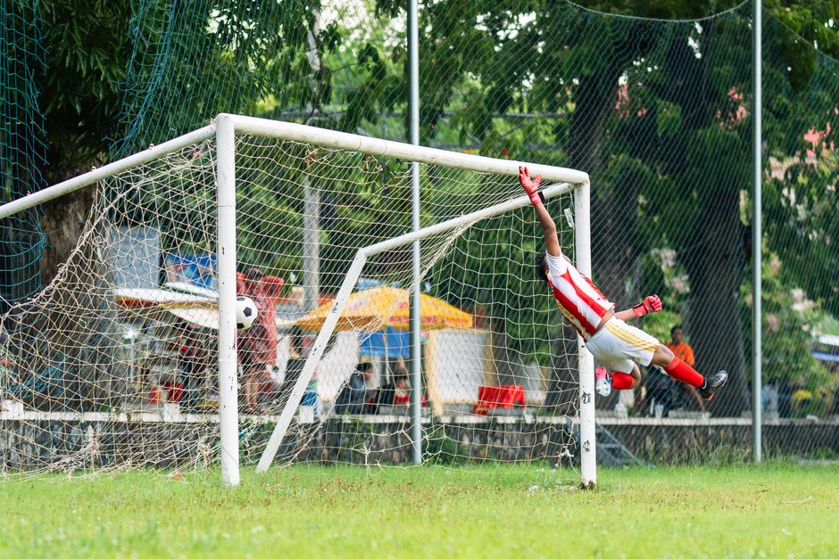 Goleiro fazendo uma defesa em jogo de futebol ao ar livre, demonstrando ação esportiva e habilidade na defesa do gol.