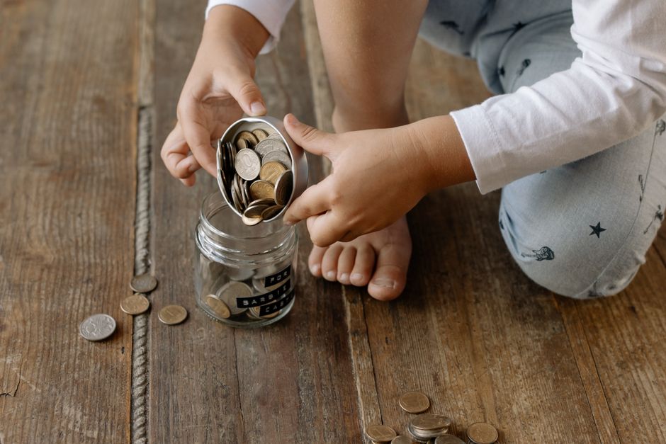 Criança colocando moedas em um cofrinho de vidro, simbolizando economia e planejamento financeiro desde cedo.