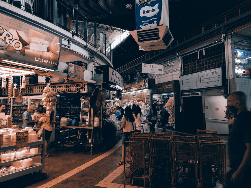 Imagem de um mercado coberto movimentado, com várias pessoas fazendo compras e explorando diferentes tendas e lojas, refletindo a dinâmica do comércio local.