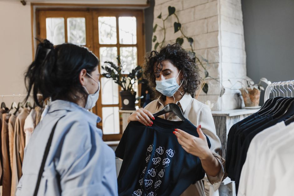 Duas mulheres usando máscaras durante uma conversa na loja de roupas, com uma delas exibindo uma blusa de manga longa com estampa de abacaxis, destacando compras e moda consciente.