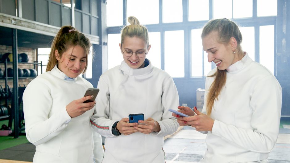 Três mulheres treinadas, sorridentes, usando roupas esportivas, checando seus smartphones em um ambiente de academia ou espaço de treinamentos ao fundo.