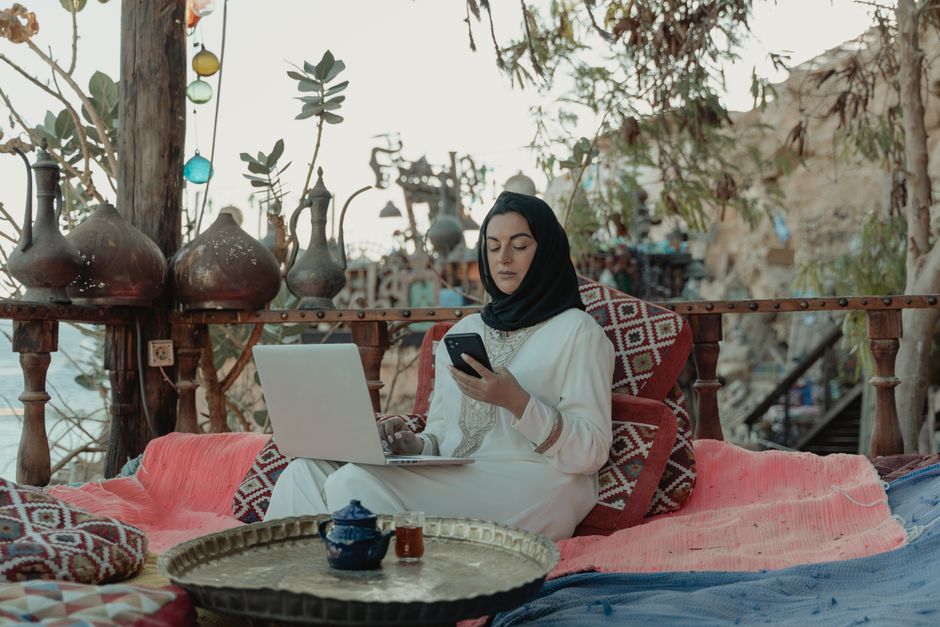 Mulher usando hijab trabalhando com laptop e celular em ambiente ao ar livre, misturando tecnologia e cultura em um espaço relaxante.