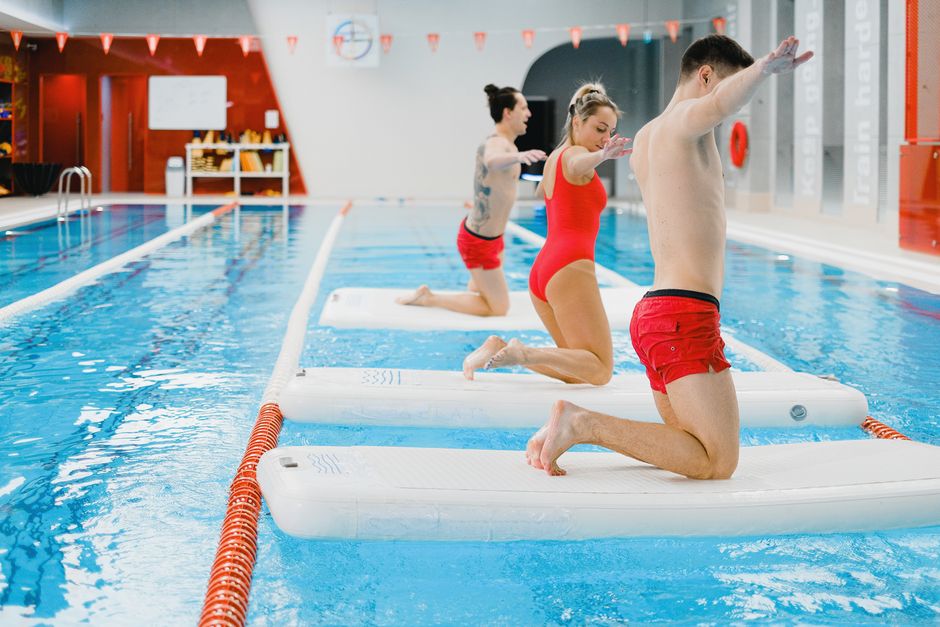 Aula de natação indoor com três adultos praticando alongamento nos tapetes flutuantes na piscina.