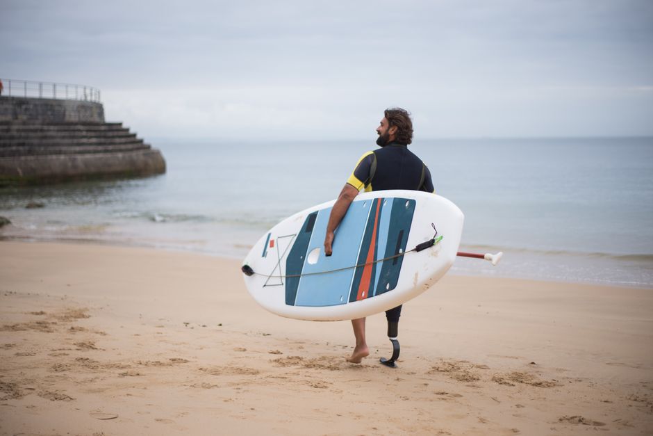 Homem com prótese na perna segurando uma prancha de surfe na praia, preparando-se para surfear em um dia nublado.