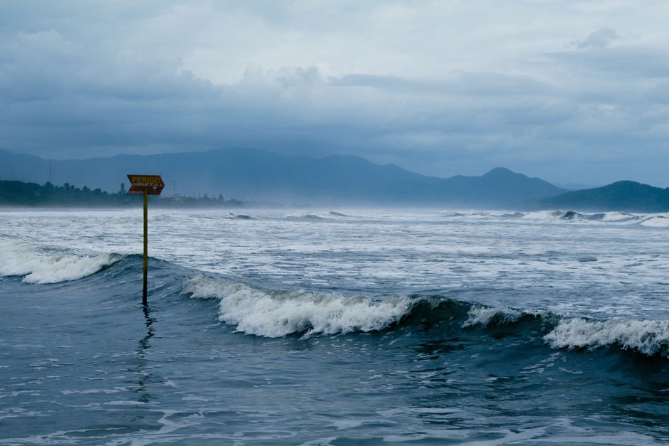 Paisagem de uma praia com ondas e uma sinalização de perigo, representando os riscos do mar e a importância da segurança costeira.