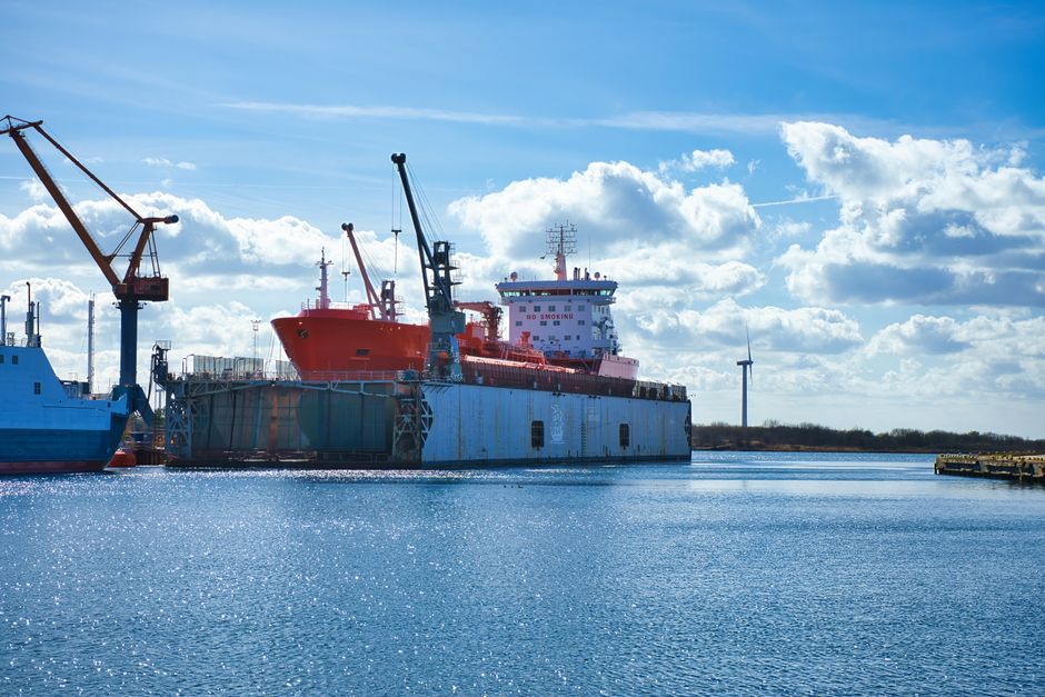 Imagem de um navio de transporte de carga atracado em um píer sob um céu parcialmente nublado, representando temas de logística e transporte marítimo.