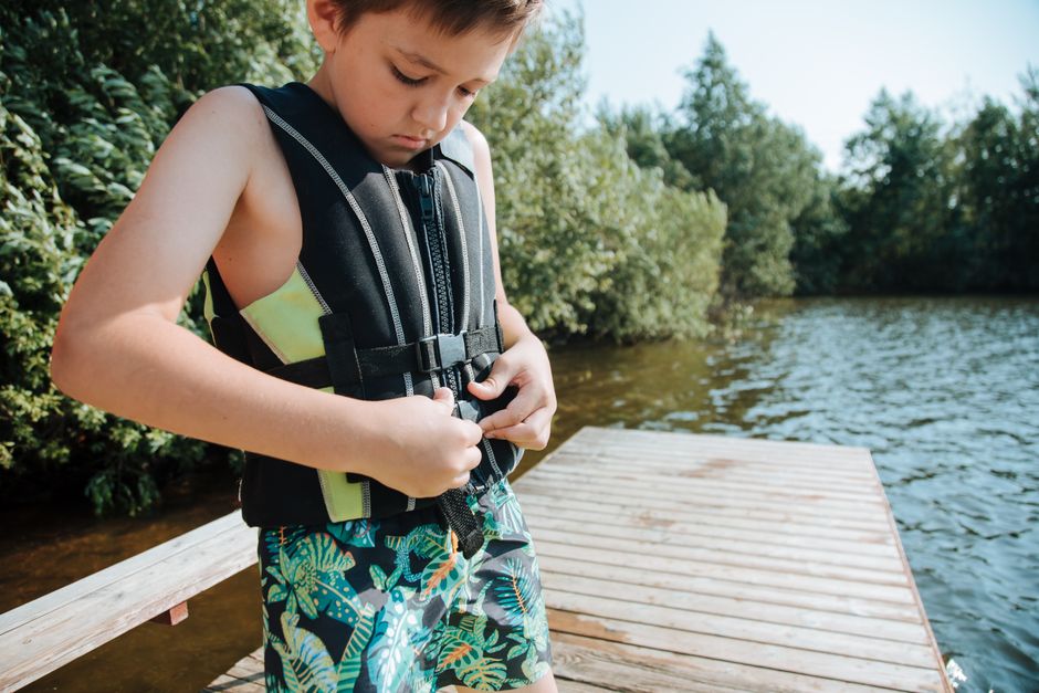 Criança vestida com colete salva-vidas, preparando-se para nadar ou explorar uma área próxima ao rio, em um ambiente ao ar livre.