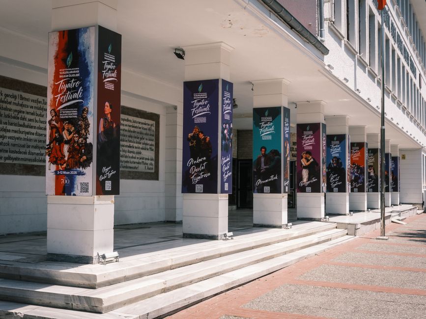 Imagem da fachada de um teatro com painéis de divulgação de um festival de teatro, exibindo figuras de performers e informações do evento.