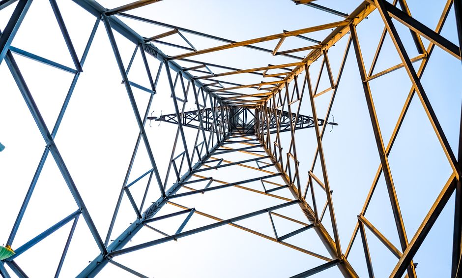 Perspectiva de uma torre de transmissão vista de baixo para cima, destacando sua estrutura metálica e o céu ao fundo, simbolizando infraestrutura e conectividade.