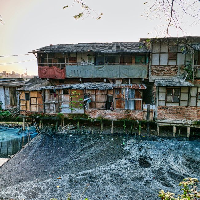 Imagem de uma habitação de construção precária próxima a um corpo d'água contaminado, refletindo questões de moradia social e meio ambiente.