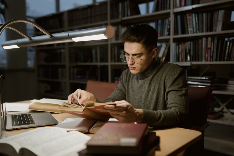 Estudante focado estudando em uma biblioteca com várias estantes de livros ao fundo, sob iluminação de mesa para pesquisa ou estudo noturno.
