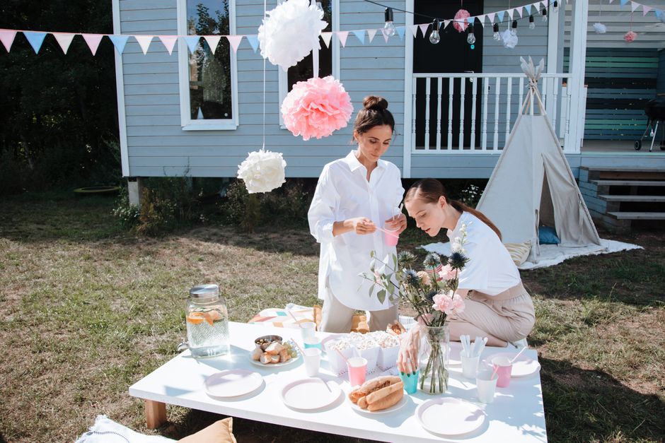 Duas mulheres preparando uma festa ao ar livre em um jardim, com uma mesa decorada para um evento descontraído e festivo.