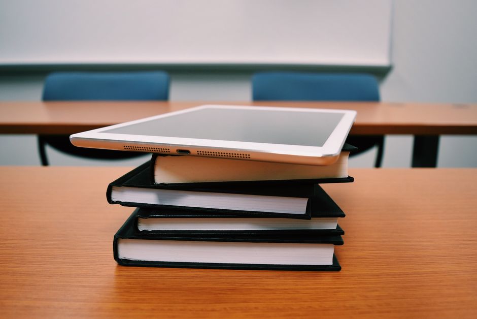 Imagem que representa estudos e recursos pedagógicos, com tablets e livros empilhados em uma sala de aula ou ambiente de estudo.