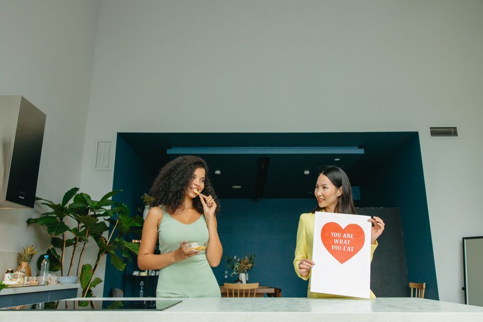 Duas mulheres em uma conversa descontraída na cozinha, com uma delas segurando uma placa que diz 'You Are What You Eat', ilustrando temas de alimentação consciente e bem-estar.