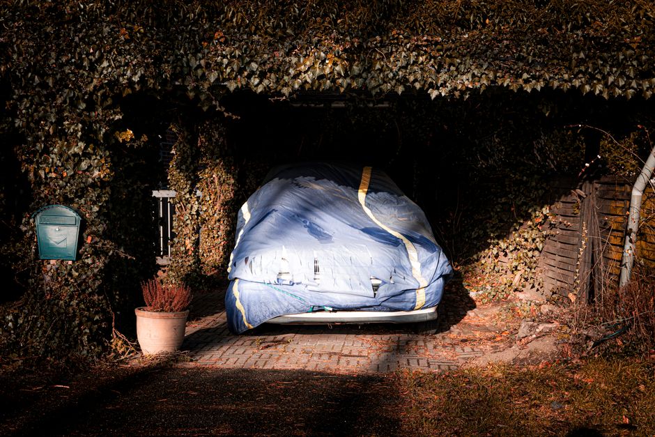 Imagem de uma garagem coberta com um carro parcialmente protegido por um cobertor, cercada por elementos de um ambiente residencial ao entardecer.