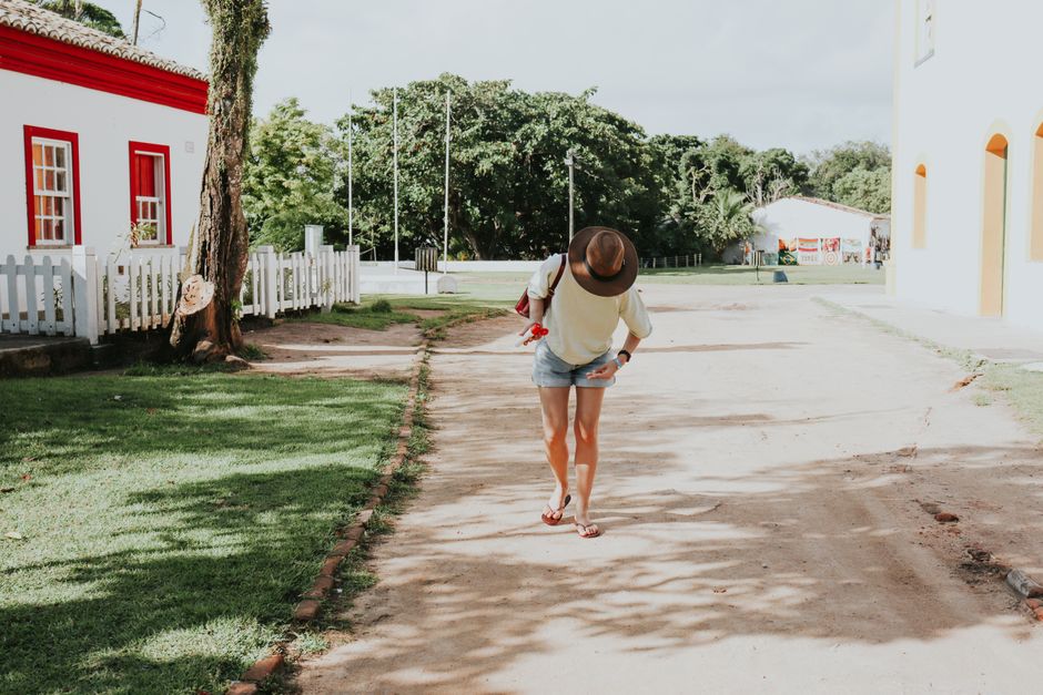 Pessoa caminhando ao ar livre em um ambiente de vila ou cidade com casas coloridas e árvores ao fundo.