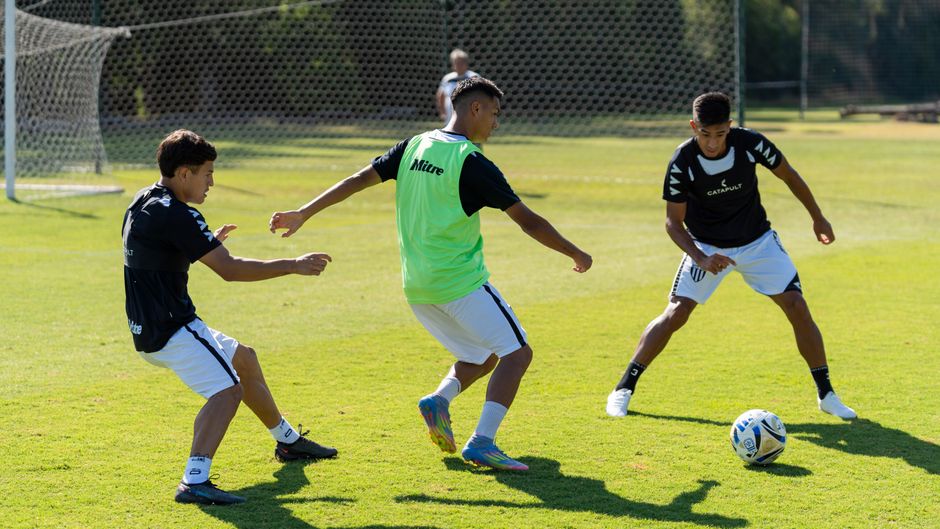 Três jovens jogando futebol em campo ao ar livre durante o dia, demonstrando prática esportiva e convivência em equipe.