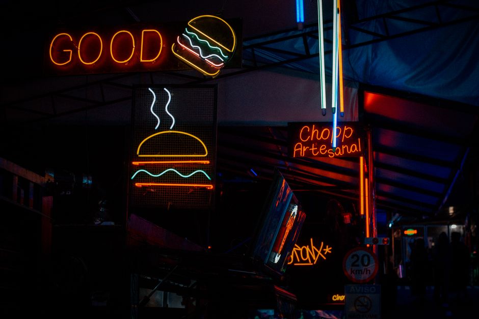 Imagem de neon com letreiros promocionais de um food truck ou feira, destacando hamburgueria artesanal e culinária de rua, em ambiente noturno.