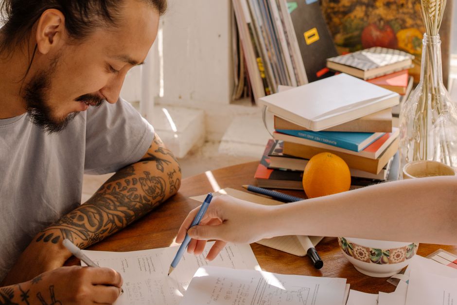 Pessoa escrevendo em documentos enquanto estuda ou trabalha em um ambiente doméstico, com livros e objetos de leitura ao redor.