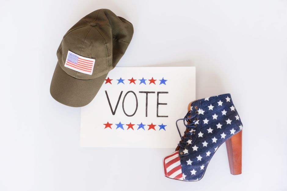 Foto com tema de votação nos Estados Unidos, apresentando um boné de campanha e uma bota com estampa da bandeira americana ao lado de uma placa com a palavra 'VOTE'.