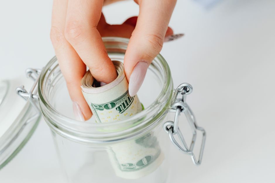 Mão colocando dinheiro em um pote de vidro, simbolizando economia ou poupança financeira.