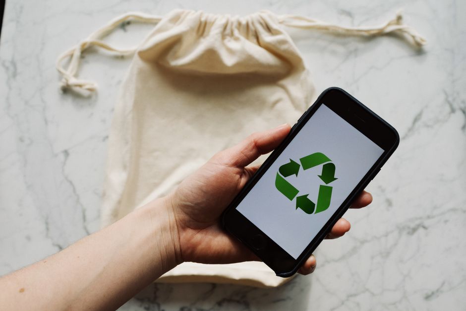 Pessoa segurando smartphone exibindo símbolo de reciclagem, com uma sacola de algodão ao fundo, simbolizando sustentabilidade e consumo consciente.