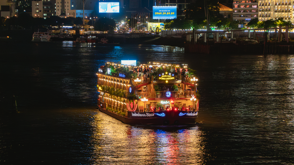 Imagem de um barco decorado e iluminado navegando por um rio à noite, com a cidade ao fundo brilhando com luzes.