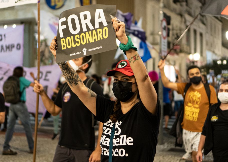 Manifestantes protestando contra o governo, com destaque para uma mulher segurando cartaz de oposição ao presidente Bolsonaro, em uma manifestação urbana noturna.