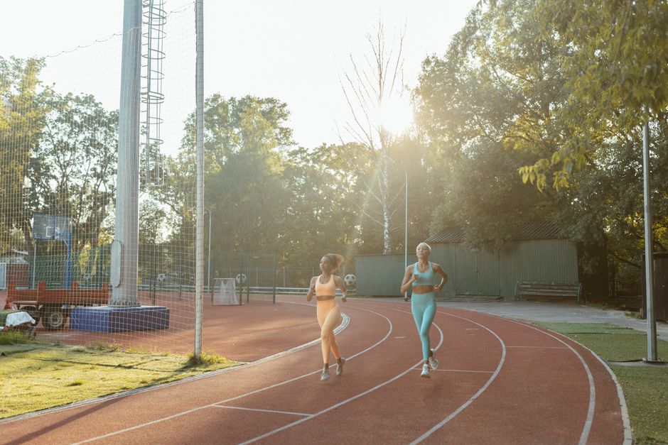 Duas mulheres praticando corrida ao ar livre em uma pista de atletismo ao entardecer, com árvores e iluminação natural ao fundo.