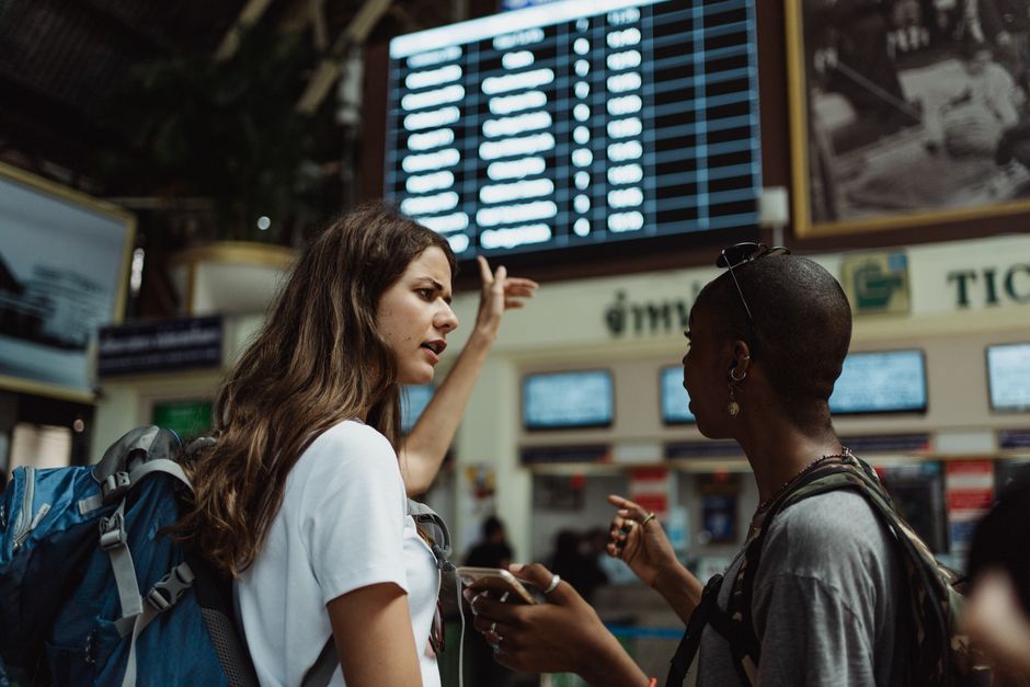 Duas viajantes discutindo ou explicando informações em um aeroporto, com um painel de informações de voos ao fundo.