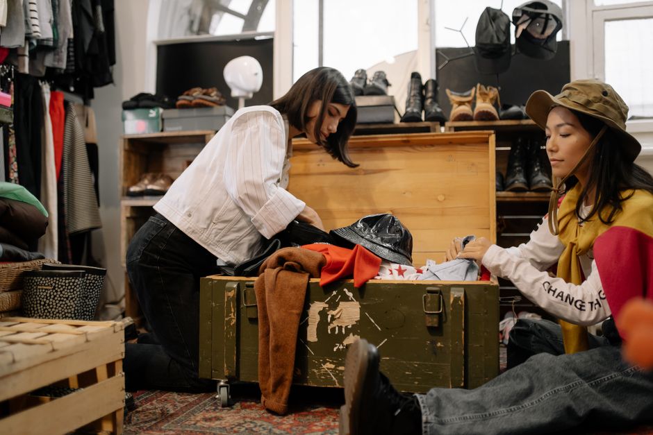 Duas mulheres explorando roupas em uma loja de segunda mão ou brechó, focando na pesquisa de peças únicas e sustentáveis.