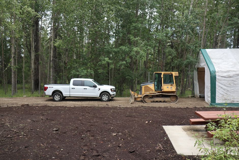 Imagem de uma área de construção ao lado de uma floresta, com um caminhonete branco e uma máquina de escavação, indicando um projeto de manutenção ou expansão ao ar livre.