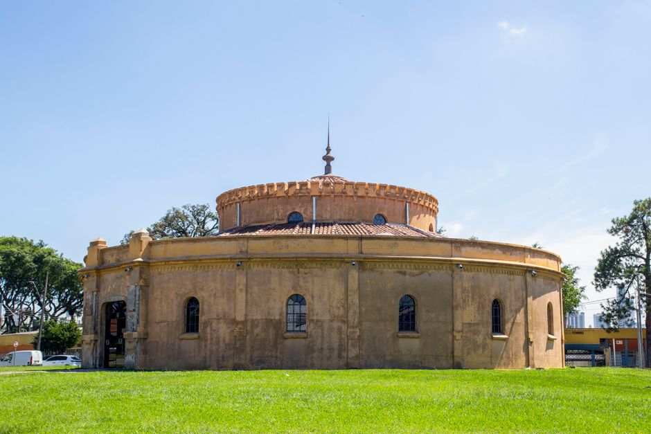 Imagem de um edifício histórico de arquitetura circular, com detalhes antigos, situado em um espaço aberto com grama e árvores ao entorno, sob céu claro.