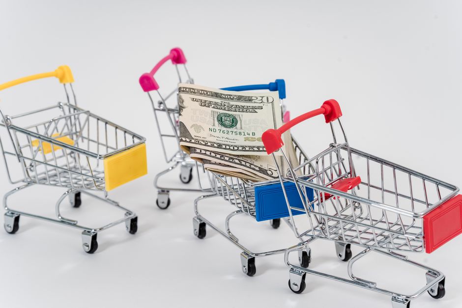Imagem de carrinhos de compras de brinquedo, um deles carregando dinheiro, simbolizando temas de economia, consumo ou finanças pessoais.