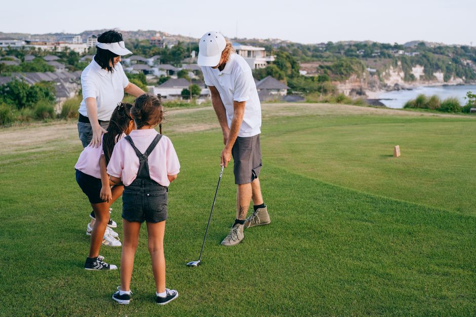 Grupo de pessoas aprendendo a jogar golfe ao ar livre em um campo com vista para natureza e rio ao fundo.