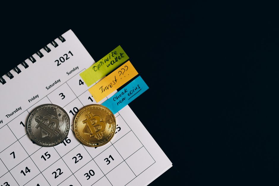 Imagem de um calendário de 2021 com duas moedas de bitcoin sobreposto, acompanhado de notas adesivas com anotações de planejamento financeiro e investimentos, sugerindo foco em estratégias de criptomoedas e gestão financeira.