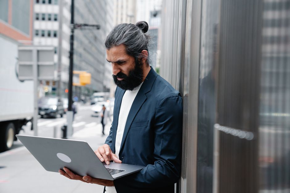 Homem com aparência profissional usando laptop na rua de uma cidade moderna, transmitindo a ideia de trabalho urbano ou empreendedorismo remoto.