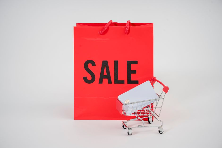 Imagem de uma sacola de compras vermelha com a palavra 'SALE' escrita, acompanhada de um carrinho de compras pequeno com cartão de pagamento, representando promoções e descontos no comércio varejista.