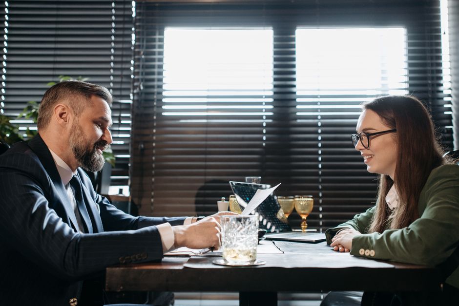 Imagem de uma reunião de negócios entre um homem de terno e uma mulher com óculos, em um ambiente corporativo com janelas de persianas ao fundo.