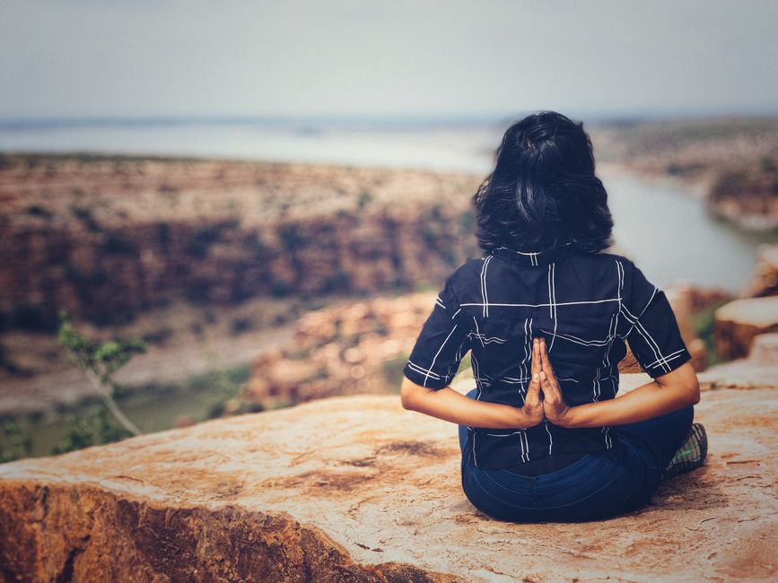 Mulher praticando meditação ou yoga ao ar livre com uma vista de cânion e rio ao fundo, transmitindo tranquilidade e conexão com a natureza.