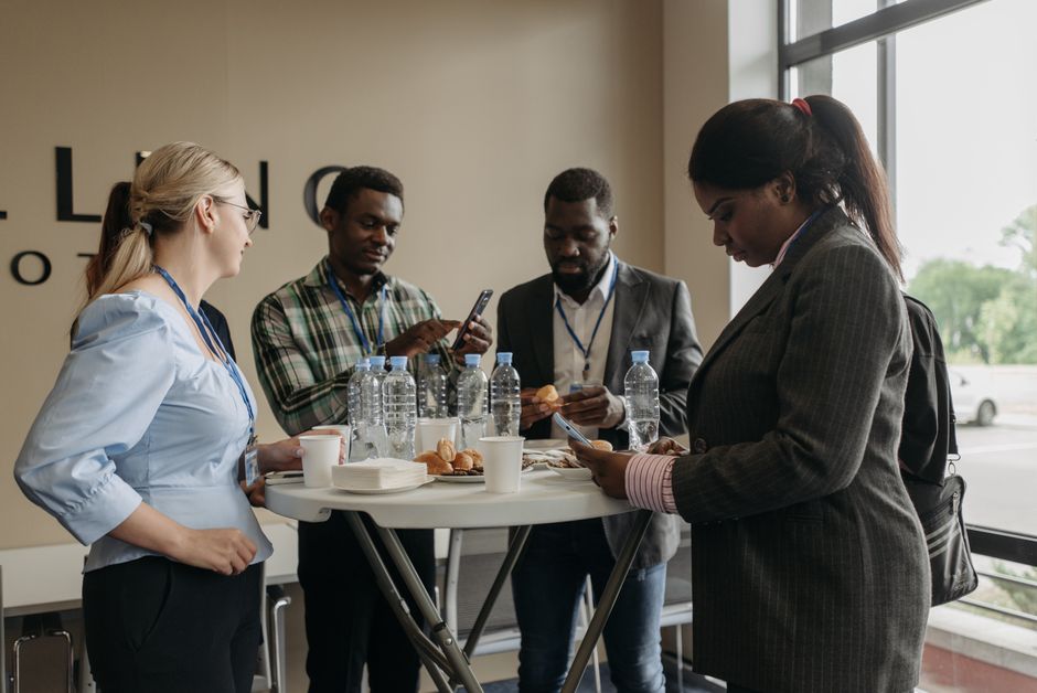 Grupo de profissionais em uma pausa de networking durante evento corporativo, usando celulares e interagindo entre si.