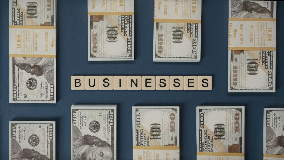 Imagem de várias pilhas de notas de dinheiro ao redor da palavra 'BUSINESSES' em blocos, representando temas financeiros e empresariais.