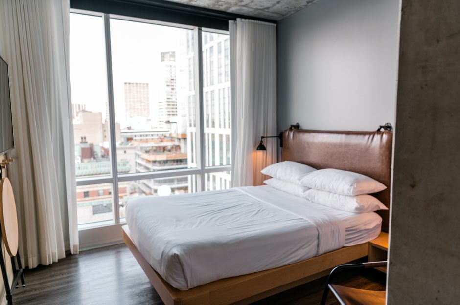 Quarto de hotel com vista urbana, destaque para a cama e janelas amplas que proporcionam luminosidade natural.