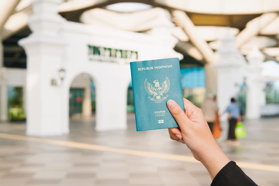 Pessoa segurando um passaporte da Indonésia em frente a uma entrada de aeroporto, simbolizando viagens internacionais.