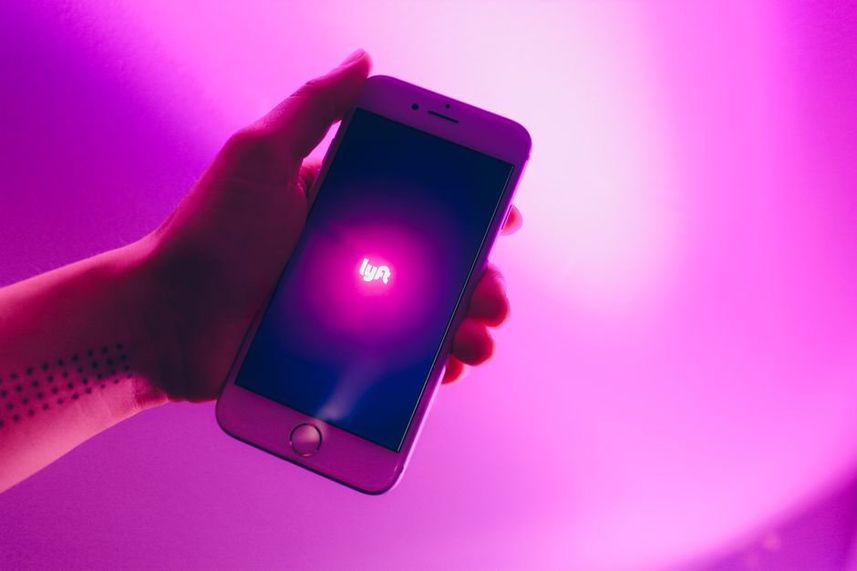 Imagem de uma mão segurando um smartphone com o logo do Uber Lyft na tela, em um ambiente com iluminação rosa e roxa, representando transporte por aplicativo.