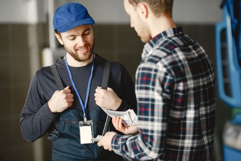 Dois homens conversando em um ambiente industrial, um deles sorrindo e segurando dinheiro, enquanto o outro oferece uma encomenda ou documento, destacando uma interação comercial ou de negócios.